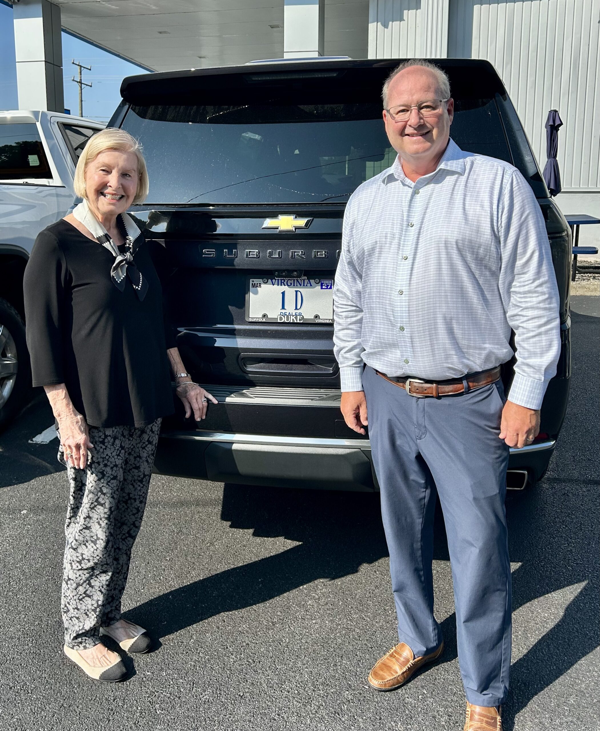 Eley Duke III and his mother, Lydia Duke, standing behind a black Chevrolet Suburban with a Virginia “1D” dealer license plate at Duke Chevrolet GMC in Suffolk
